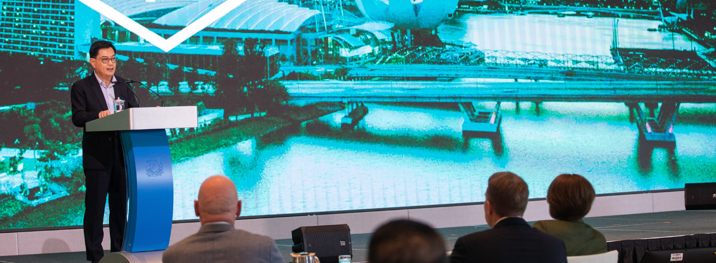 Man speaks at podium with Singapore flag; audience views building reflection on screen.